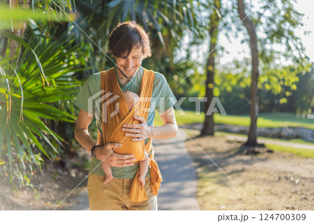 Father holding his little baby in a yellow sling in the park. Warm and loving family moment. Babywearing, fatherhood, and parent-child bonding concept Father holding his little baby in a yellow sling in the park. Warm and loving family moment. Babywearing, fatherhood, and parent-child bonding concept 124700309