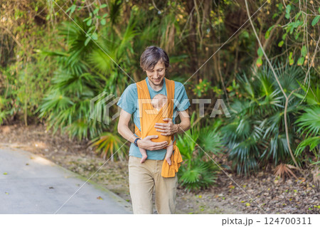 Father holding his little baby in a yellow sling in the park. Warm and loving family moment. Babywearing, fatherhood, and parent-child bonding concept 124700311