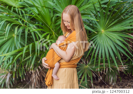 Mother holding her little baby in a yellow sling in the park. Warm and loving family moment. Babywearing, parenting, and mother-child bonding concept Mother holding her little baby in a yellow sling in the park. Warm and loving family moment. Babywearing, parenting, and mother-child bonding concept 124700312