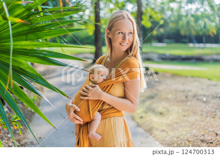 Mother holding her little baby in a yellow sling in the park. Warm and loving family moment. Babywearing, parenting, and mother-child bonding concept Mother holding her little baby in a yellow sling in the park. Warm and loving family moment. Babywearing, parenting, and mother-child bonding concept 124700313