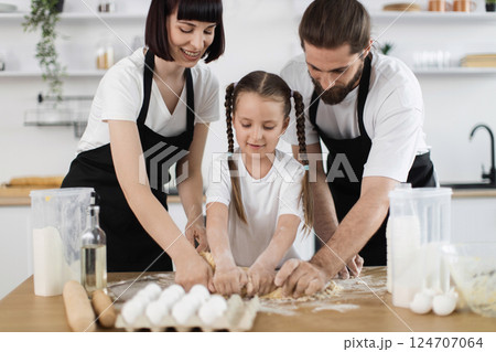 Caucasian family, including parents and young girl, participate in cooking activity, kneading dough attentively wearing white shirts and black aprons, symbolizing teamwork 124707064