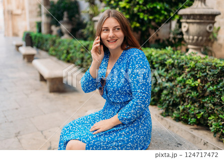 Beautiful stylish woman in trendy wear spending time on street using smartphone. Happy girl in dress talking on the mobile phone. Urban lifestyle concept, old town 124717472