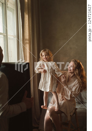Mother plays with her daughter in a warm and cozy room filled with natural light while father captures the joyful moment during a family photoshoot Mother plays with her daughter in a warm and cozy room filled with natural light while father captures the joyful moment during a family photoshoot 124721627