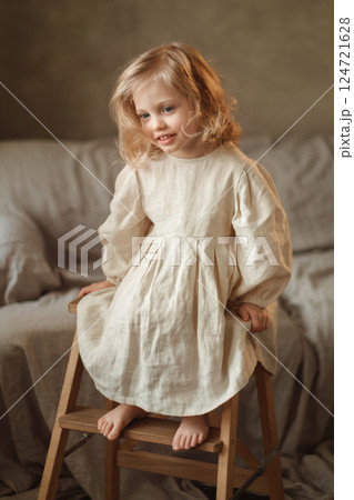 Young girl sitting on a wooden stool wearing a light-colored dress in a cozy indoor setting with soft textiles and warm lighting 124721628