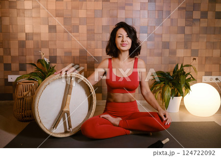 A woman in a red tracksuit sits with a drum in a gymnasium. Kalimeditation class 124722092