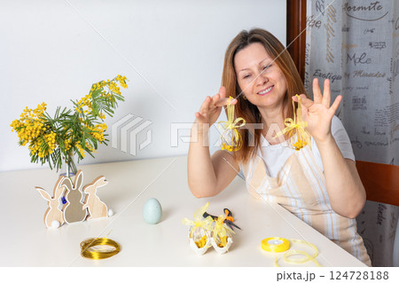 Smiling woman making handmade Easter egg wire ornaments with mimosa and yellow ribbon at a festive spring table. Creative holiday crafts and DIY Easter decoration. 124728188