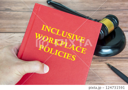 Hand holding Inclusive Workplace Policies book with gavel on a wooden table background 124731591