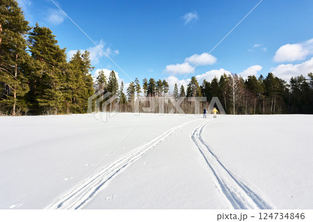 Distant figures ski across snow covered field, one wearing bright yellow clothing while other follows, both moving toward distant treeline on sunny day. Distant figures ski across snow covered field, one wearing bright yellow clothing while other follows, both moving toward distant treeline on sunny day. 124734846