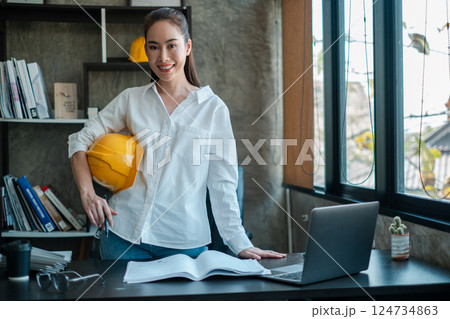 Female architect in a modern office, holding a safety helmet, with blueprints and a laptop on the desk, smiling confidently. 124734863