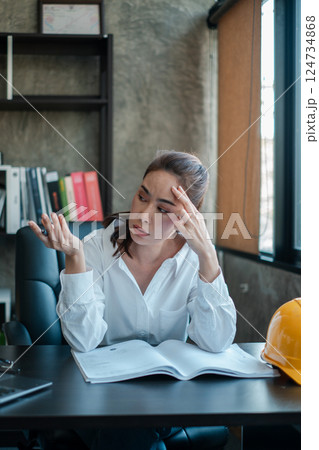 A businesswoman in a white shirt sits at a desk, appearing thoughtful, with an open book and a construction helmet nearby. A businesswoman in a white shirt sits at a desk, appearing thoughtful, with an open book and a construction helmet nearby. 124734868