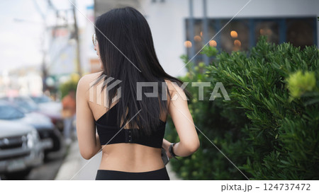 Young woman with long dark hair walking along a street in urban setting, wearing black outfit and showcasing a relaxed lifestyle in a vibrant city environment 124737472