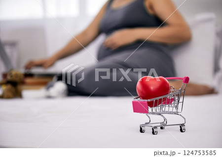 Mini shopping cart with red heart in focus on bed with pregnant woman in gray dress using laptop in background; love care online shopping and motherhood 124753585