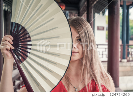 Young woman holds a decorative fan near her face in outdoor area with traditional architecture in the background. Young woman holds a decorative fan near her face in outdoor area with traditional architecture in the background. 124754269