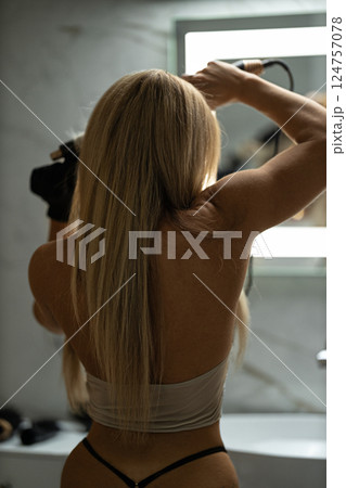 woman long hair stands in front of mirror using blow dryer style her hair. modern bathroom setting features marble and bright lighting creating fresh atmosphere. woman long hair stands in front of mirror using blow dryer style her hair. modern bathroom setting features marble and bright lighting creating fresh atmosphere. 124757078