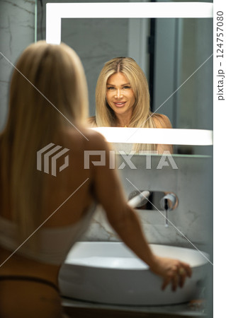 woman stands stylish bathroom sink smiling her reflection in illuminated mirror showcasing her long blonde hair and relaxed morning vibe. woman stands stylish bathroom sink smiling her reflection in illuminated mirror showcasing her long blonde hair and relaxed morning vibe. 124757080