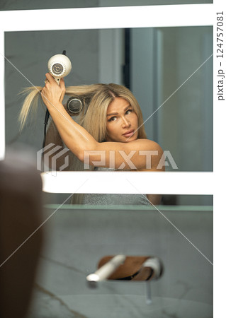 In contemporary bathroom woman long blonde hair is styling her locks using hair dryer. mirror reflects her focused expression as she enhances her look. In contemporary bathroom woman long blonde hair is styling her locks using hair dryer. mirror reflects her focused expression as she enhances her look. 124757081