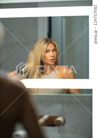 In chic bathroom woman is using round brush style her long hair while gazing her reflection in well lit mirror. Soft colors create calming atmosphere. In chic bathroom woman is using round brush style her long hair while gazing her reflection in well lit mirror. Soft colors create calming atmosphere. 124757082