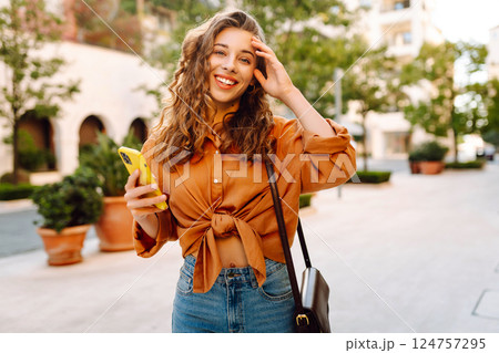 Portrait of a young woman holding a phone in her hands on a city avenue, on a sunny day. 124757295
