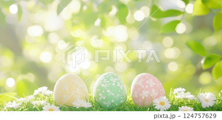 Three colorful Easter eggs with delicate floral patterns resting on fresh green grass, surrounded by tiny white daisies. Sunlight filters through vibrant leaves, creating a dreamy bokeh background 124757629