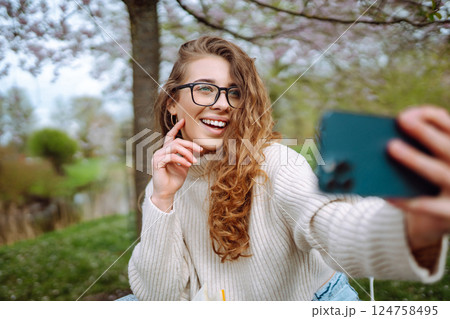 Young woman takes selfie against background of flowering trees in park. Concept of relaxation, fun. 124758495