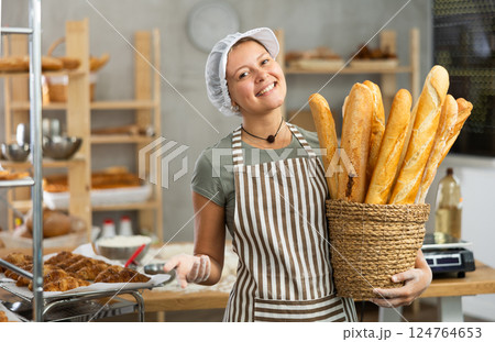 Bakery female seller stands proudly in front of fresh bread holding basket of freshly baked baguettes in kitchen 124764653