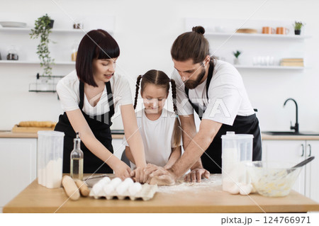 Caucasian family cooking in kitchen having fun together. Mother, father and daughter wearing aprons working with dough, kitchen filled with flour and ingredients on table, enjoying happy bonding time. 124766971