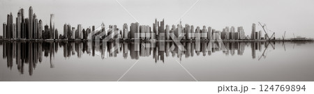 Monochrome city skyline with tall skyscrapers reflecting on calm water under a cloudy sky, Dubai Marina bay UAE 124769894