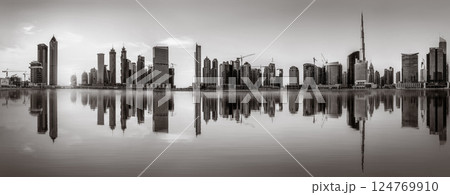 Monochrome city skyline with tall skyscrapers reflecting on calm water under a cloudy sky, Dubai Marina bay UAE 124769910