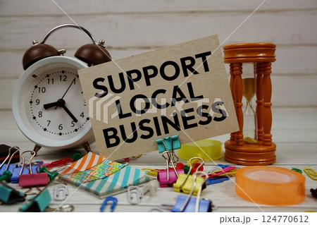 Support Local Business text on paper card with alarm clock and stationary on wooden background 124770612