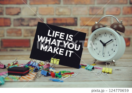 Life Is What You Make It text on paper card with alarm clock and stationary on wooden background 124770659