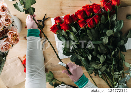 Florist trimming red rose stems by hand with a knife beside fresh bouquets and greenery. Bouquet assembly, floristry craftsmanship, slow production, floral detailing, hands-on work Florist trimming red rose stems by hand with a knife beside fresh bouquets and greenery. Bouquet assembly, floristry craftsmanship, slow production, floral detailing, hands-on work 124772552