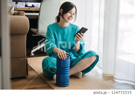 Young woman sitting on a cork mat using a smartphone beside a foam roller. Wellness meets tech, digital self-care routines, guided mobility, smart training 124772555