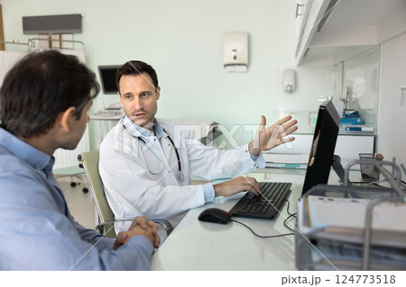 Doctor sits in workplace with computer, explaining information to patient Doctor sits in workplace with computer, explaining information to patient 124773518