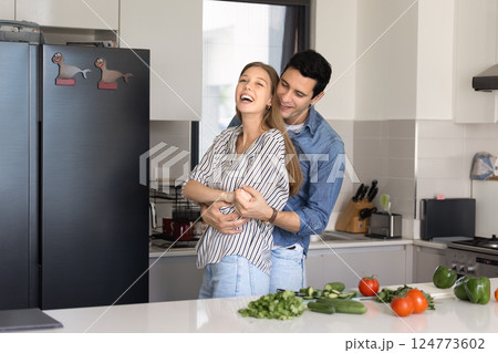 Cheerful couple hugging, distracted from dinner preparation in modern kitchen 124773602