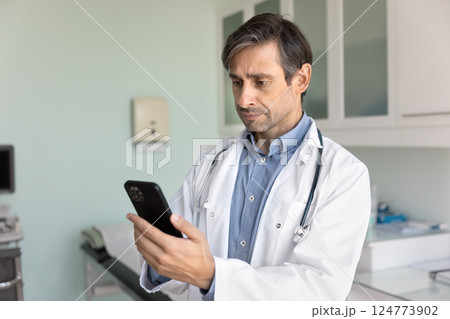 Physician in coat using smartphone in consulting room 124773902