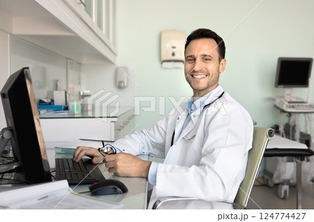 Portrait of smiling professional GP sit at desk with computer Portrait of smiling professional GP sit at desk with computer 124774427