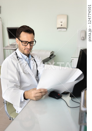 Doctor wearing glasses and white coat attentively reviewing medical documents Doctor wearing glasses and white coat attentively reviewing medical documents 124774501