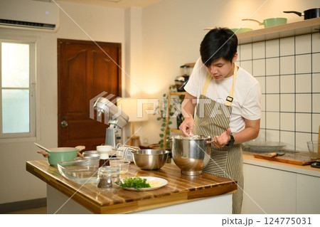 Young asian man preparing whipped cream in stainless steel bowl, using whisk in a bright kitchen 124775031