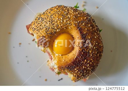 Close-up of bitten bagel with egg, ham, cheese and fresh greens. Half-eaten breakfast sandwich on plate, top view Close-up of bitten bagel with egg, ham, cheese and fresh greens. Half-eaten breakfast sandwich on plate, top view 124775512