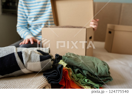 Woman packs donation box with clothes at home. Person places warm sweater into cardboard box. Preparing used clothing for recycling or second hand. Concept of charity and humanitarian aid Woman packs donation box with clothes at home. Person places warm sweater into cardboard box. Preparing used clothing for recycling or second hand. Concept of charity and humanitarian aid 124775540
