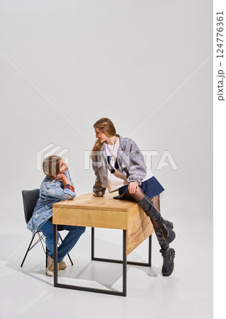 Two students interact near wooden desk against plain background. Boy sits on black chair, looking up at girl, who leans on desk. Two students interact near wooden desk against plain background. Boy sits on black chair, looking up at girl, who leans on desk. 124776361
