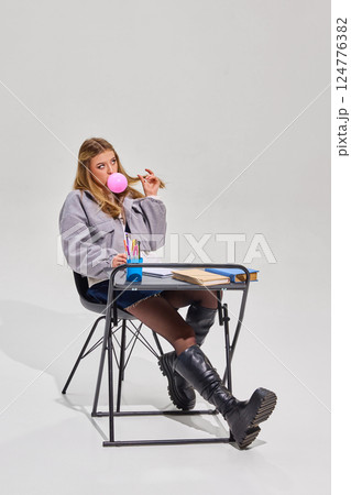 Blonde girl in grey jacket and black boots blows pink bubblegum bubble while sitting at desk with books, notebook, and cup of colorful pens. Blonde girl in grey jacket and black boots blows pink bubblegum bubble while sitting at desk with books, notebook, and cup of colorful pens. 124776382