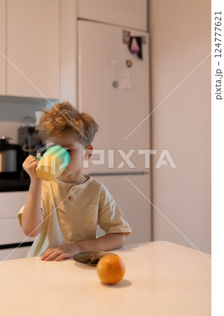 Young boy enjoying a warm drink at a kitchen table in the morning with a fresh orange nearby Young boy enjoying a warm drink at a kitchen table in the morning with a fresh orange nearby 124777621