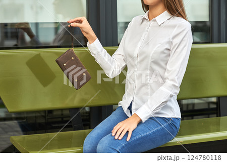 Woman in casual attire holding brown wallet on park bench, stylish and modern accessory concept 124780118