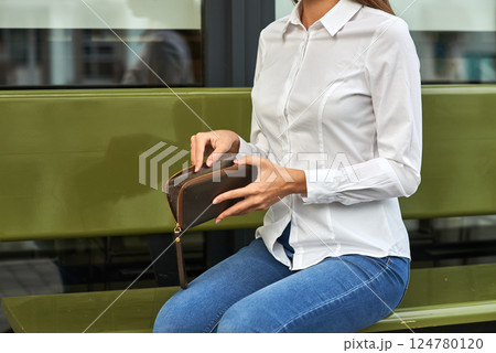 Woman in white shirt, jeans, holding leather wallet, sitting on green bench, outdoor, style, casual, urban lifestyle, fashion concept 124780120