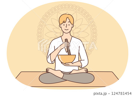 Meditating man sitting in lotus position on yoga mat using bowl to perform ancient ritual 124781454