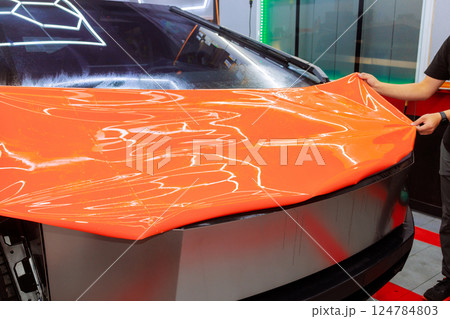 Worker carefully applies bright orange vinyl wrap to front of modern car in detailing studio. 124784803