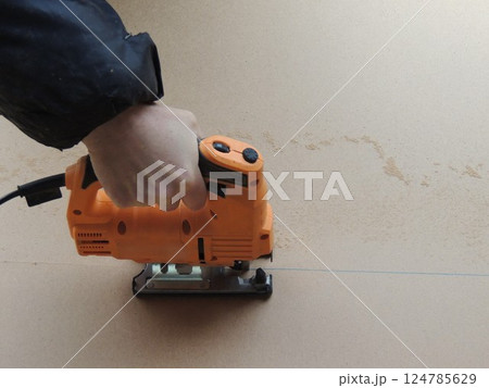 hand presses orange jigsaw and moves along marking line, cutting sheet of pressed wood into pieces, cutting sheet of plywood lengthwise with jigsaw along vertical marking line 124785629