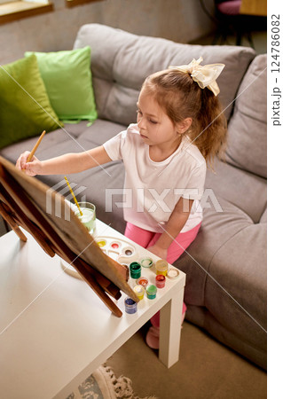 Focused girl, elementary school pupil sitting on sofa and painting, expressing creativity while learning at home. 124786082