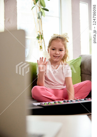 Girl, elementary school pupil sitting on sofa, smiling and waving to camera during online music lesson, sitting with keyboard on couch. Girl, elementary school pupil sitting on sofa, smiling and waving to camera during online music lesson, sitting with keyboard on couch. 124786098